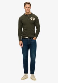 Oliven grøn langærmet henley-skjorte med rund hals og knaplukning, kombineret med mørkeblå jeans og lysebeige sneakers.