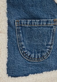 Tascone in denim con bordi arrotondati, caratterizzato da cuciture visibili in filo arancione, affiancato da un morbido rivestimento in pile color crema.