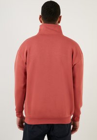 Fleece pullover in koraal, met een hoge halslijn, verlaagde schouders en ribgebreide manchetten. Gladde textuur zonder zichtbare patronen.