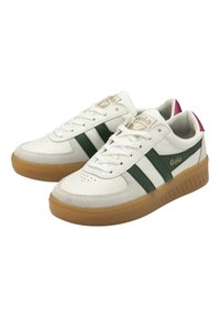 Zapatilla blanca con rayas verdes, detalles de ante y una suela de goma. Presenta un collar acolchado y cierre con cordones. Logo de Gola en el costado.