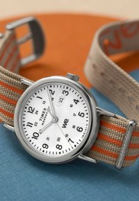 Montre en acier inoxydable argenté avec un cadran blanc, des marqueurs horaires noirs et un affichage de date. Bracelet en tissu beige avec des rayures orange.