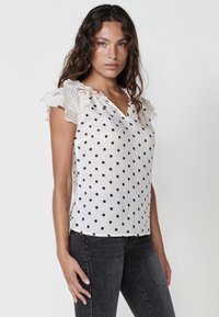 Blusa bianca con pois neri, con maniche trasparenti arricciate e scollatura a V, abbinata a jeans grigi scuri.