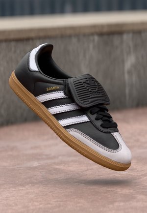 Sort-hvid Adidas Samba-sneaker med gummisål, sorte snørebånd og en foldet tunge, der viser Adidas-logoet, svævende over jorden.