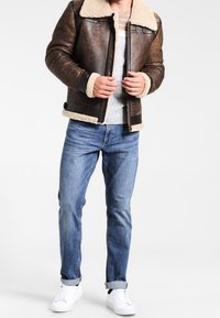 Veste en cuir marron avec col en peau lainée, fermeture éclair et poches latérales. Portée sur un haut gris à rayures horizontales et un jean bleu.