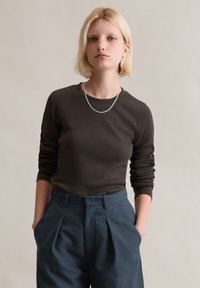 Haut à manches longues côtelé marron avec encolure ras du cou, associé à un pantalon bleu taille haute à plis. Accent de collier chaîne argentée.