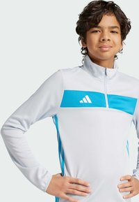 Maglia sportiva bianca a maniche lunghe con collo alto, caratterizzata da una striscia orizzontale blu e dettagli laterali blu, realizzata in un tessuto liscio ed elastico.