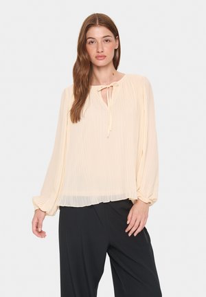 Light beige blouse with pleated texture, round neckline, and a bow tie closure. Features long, puffed sleeves and a relaxed fit.