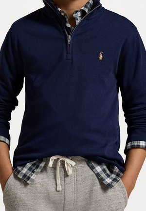 Persona vistiendo un suéter azul marino con cremallera parcial sobre una camisa de cuadros, con pantalones de chándal grises con cordón, manos en los bolsillos, torso visible, sin mostrar el rostro.