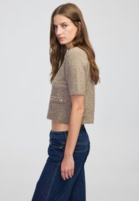 Kobieta z długimi brązowymi włosami, ubrana w beżowy sweter z krótkim rękawem i ciemnoniebieskie dżinsy, stojąca bokiem na jednolitym tle.