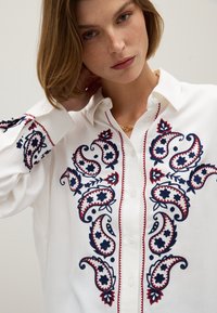 Camicia bianca con bottoni caratterizzata da un ricamo intricato paisley blu navy e rosso, con accenti floreali lungo la patta e i polsini delle maniche.