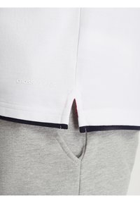 Biała koszulka polo z granatowym wykończeniem rękawów i dolnego brzegu; z logo "CROSS JEANS" w białym kolorze, w zestawieniu z jasnoszarymi spodniami dresowymi.