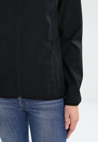 Black zip-up jacket made of smooth fabric, featuring a high collar and side zippered pockets. Worn with blue jeans.