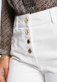 Los pantalones vaqueros blancos de tiro alto cuentan con acentos de botones dorados, una tela suave y texturizada, y bolsillos en las costuras laterales. La cinturilla es plana y tiene un acabado nítido.