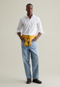 Camisa blanca de botones, suéter de punto amarillo atado a la cintura, jeans baggy de color azul claro, zapatos marrones sin cordones, de pie sobre un fondo sencillo.