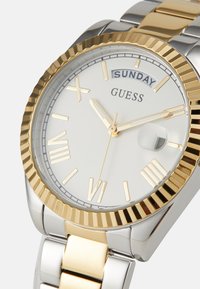 Metal watch with a two-tone silver and gold band. White face features gold Roman numerals, gold hands, and a date window beneath "SUNDAY."