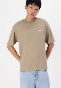 Camiseta de manga corta en tono marrón, hecha de algodón suave, con "Club1990" en texto blanco en el lado izquierdo, combinada con jeans de color azul claro.