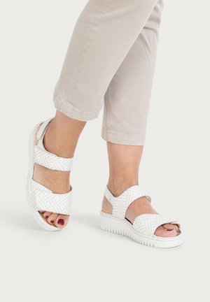 Feet wearing white patterned open-toe sandals with adjustable straps, paired with light beige cropped pants on a neutral background.