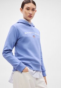 Sudadera con capucha azul claro con texto blanco y un pequeño emblema. Lleva una camiseta blanca con rayas azules debajo. Tejido suave, ajuste holgado.