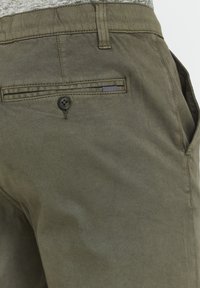 Pantalons vert olive avec une texture lisse, une poche arrière boutonnée et un accent à rayures horizontales sur la poche. Design décontracté.