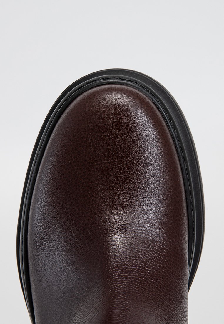 Nahaufnahme der abgerundeten Spitze eines polierten, dunkelbraunen Lederstiefels mit schwarzer Sohle und detaillierter Nahtführung.