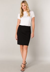 Weißes T-Shirt mit kurzen Ärmeln, kombiniert mit einem knielangen schwarzen Bleistiftrock und schwarzen, Riemchen hohen Sandalen mit Absatz. Schlichtes und figurbetontes Design.