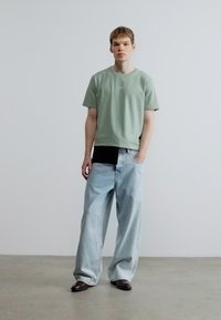 Light green short-sleeve t-shirt with small logo, paired with loose light blue jeans. Model stands with hands in pockets, wearing dark shoes.