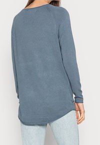 Langärmliger, hellblauer Pullover mit abgerundetem Saum, gerippter Textur und Nahtdetails an den Schultern. Getragen mit hellen Jeans.