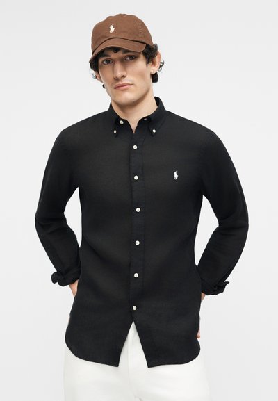 Joven con gorra de béisbol marrón, camisa negra abotonada con logo blanco y pantalones blancos, de pie con las manos detrás de la espalda.