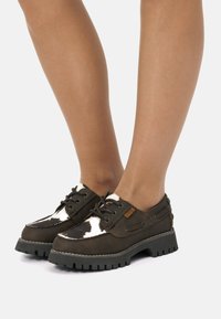 Zapatos marrones con acentos de cuero de vaca y suelas de goma oscuras. Presentan cordones, una punta redondeada y una textura resistente.