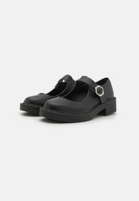 Black leather Mary Jane shoes with a round toe, adjustable strap, and a low block heel. Features contrasting stitching and a side buckle.