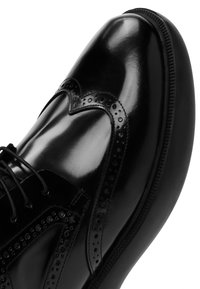 Zwarte leren schoen met glanzende afwerking, wingtipontwerp met decoratieve perforaties en ronde neus. Zwarte veters en een gestructureerde zool.