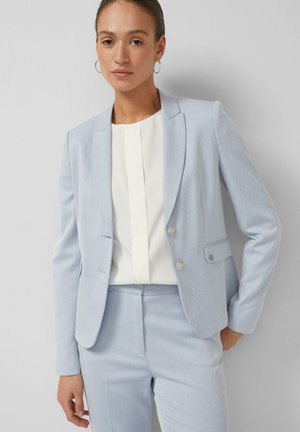 Kobieta w jasno niebieskim, dopasowanym żakiecie i pasujących spodniach oraz białej bluzce, stojąca na tle jednolitego, jasnoszarego tła.