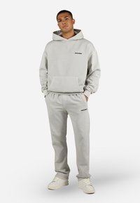 Man in een lichtgrijze hoodie en bijpassende joggingbroek met zwarte "PEGADOR" logo's, staand met handen in zijn zakken en witte sneakers.