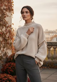 Beżowy sweter dziany z wysokim kołnierzem i oversized rękawami, zestawiony z szarymi spodniami. W tle znajdują się brązowe liście i kwiaty.