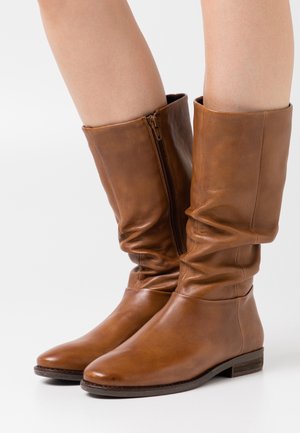 Botas de cuero marrón hasta la mitad de la pantorrilla con cremallera lateral, llevadas sobre piernas desnudas, contra un fondo blanco.