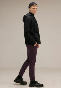 Mustat teksturoidut bleiseri huppuineen, purppurat slim-fit-housut ja mustat paksupohjaiset kengät. Malli seisoo sivuttain neutraalilla taustalla.