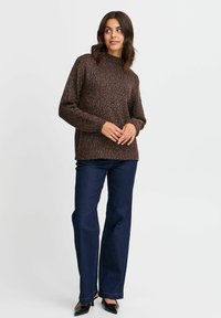 Suéter de punto marrón con manchas, de cuello alto, puños de canalé y corte relajado, combinado con pantalones de mezclilla oscuros de pierna acampanada y tacones negros.