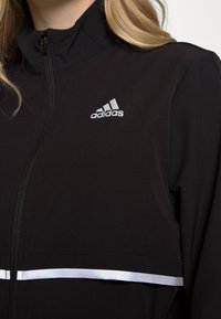Chaqueta negra con cremallera, que presenta un cuello alto, textura acanalada y una franja horizontal blanca con el logo de Adidas en el lado izquierdo del pecho.