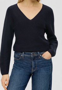 Mörkblå stickad tröja med djup V-ringning, långa ärmar och ribbad textur, kombinerad med mörkblå jeans.