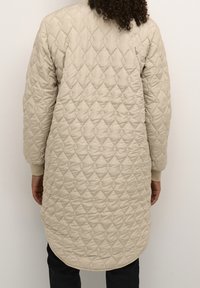 Beige värillinen pitkähihainen toppatakki, jossa on timantinmuotoinen tikkauskuvio, pyöreä helma ja resorit hihansuissa. Kevyt materiaali sileällä pintakäsittelyllä.