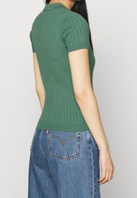 En kvinna står med ryggen mot kameran, klädd i en åtsittande grön ribbad topp och vida blå jeans med ett hjärtformat sömmönster.