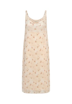 Femme portant une robe nuisette fleurie crème à fines bretelles, des boucles d'oreilles en perles et une bague, posant avec une main tenant la robe.