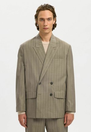 Giovane uomo con capelli castani di media lunghezza che indossa un abito beige a doppio petto a righe sottili oversize e una camicia chiara.