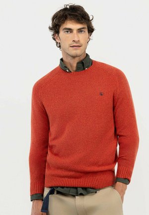 Hombre con cabello castaño rizado que lleva un suéter de punto naranja sobre una camisa abotonada verde oscuro con pantalones beige. Una mano en el bolsillo.