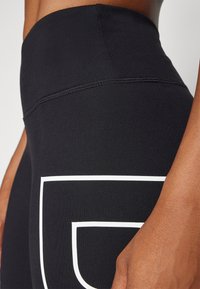 Black high-waisted leggings made of stretchy material, featuring a prominent white logo design on the thigh and flatlock seams.