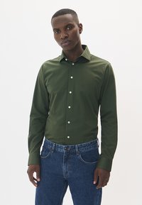 Tamnozeleni košulja s dugim rukavima i strukturiranim ovratnikom, uparen s plavim trapericama koje imaju opušteni kroj i suptilan sjaj.