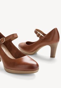 Escarpins Mary Jane en cuir marron avec bouts arrondis, brides de cheville réglables avec boucles dorées, et talons moyens empilés sur fond blanc.