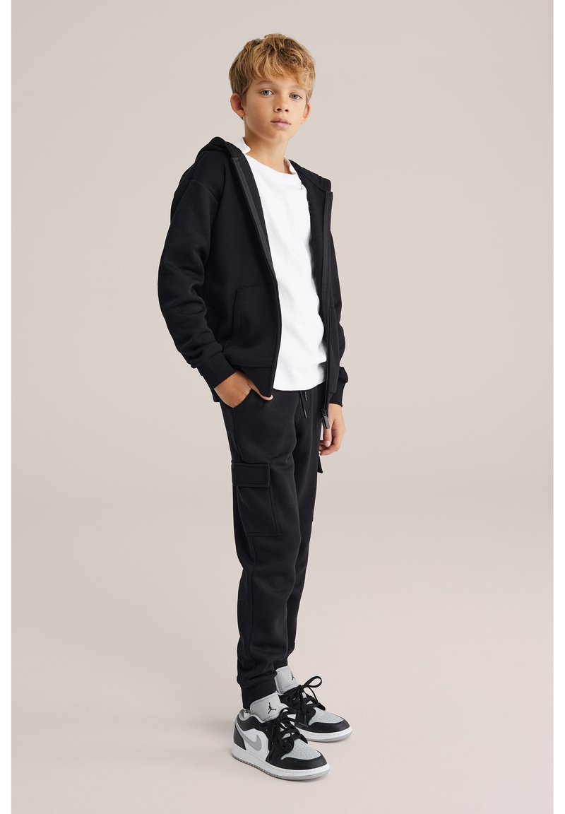 Jeune garçon debout, portant un sweat à capuche noir zippé, un t-shirt blanc, un pantalon cargo noir et des baskets noir et blanc sur un fond neutre.