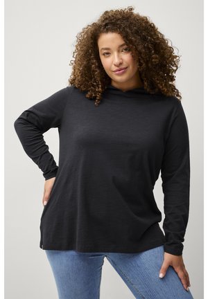 Kobieta o kręconych włosach, ubrana w czarną koszulkę z długim rękawem i niebieskie dżinsy, pozująca z jedną ręką na biodrze na jasnym tle.