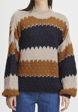 Pull en tricot épais avec de larges rayures horizontales en zigzag beige, marron et noir, porté avec un jean bleu, montrant le torse moyen et les bras.
